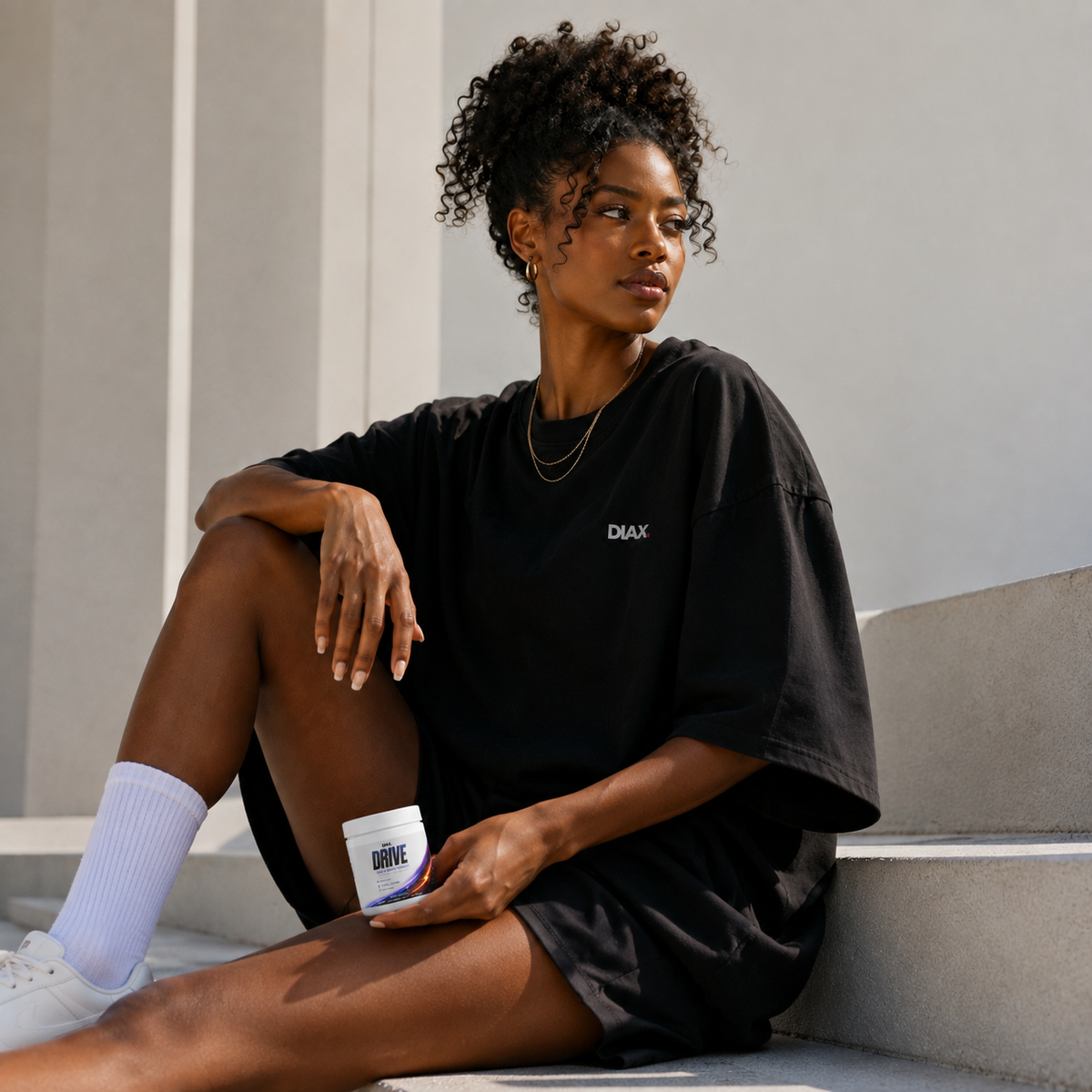 A woman sits on concrete steps wearing an oversized black t-shirt with a small white 'DMAX' logo on the chest, white socks, and white sneakers, holding a white cup with 'DRIVE' printed on it.
