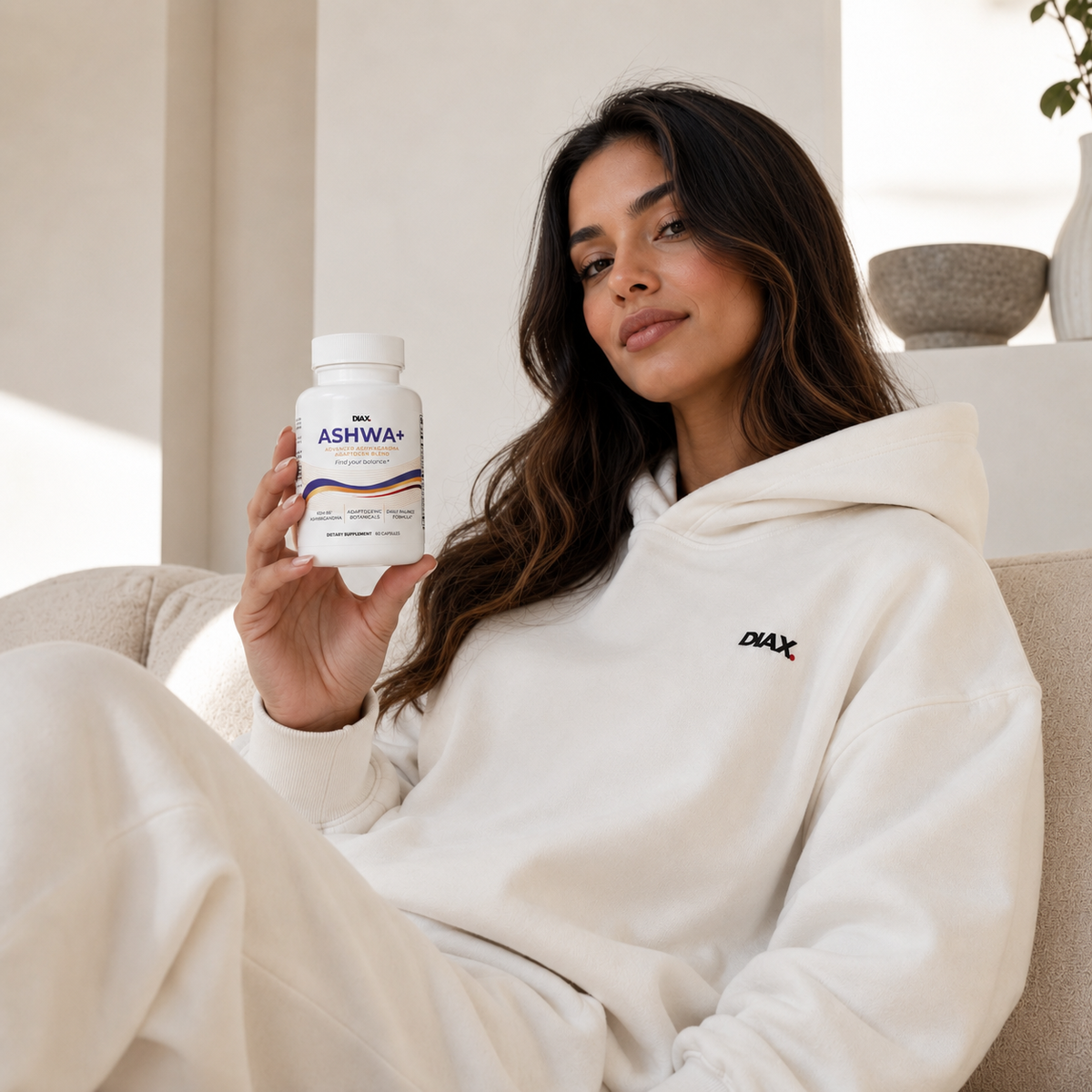 A woman in a white hoodie sits on a beige couch holding a white bottle of DMAX Ashwa+ supplements. The room has a warm, minimalist aesthetic with beige walls and soft natural light.